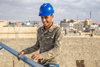 A smiling person wearing a hard hat holds construction materials.
