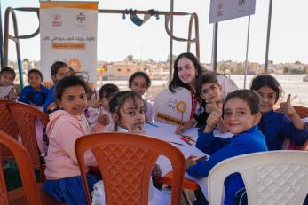 Children sitting around a table, smiling at the camera, while making art.